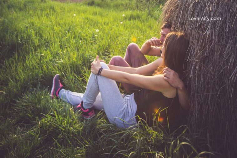 A couple sits leaning against a haystack in a grassy field, the man's arm around the woman.