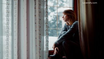 A woman in pajamas sits on a windowsill looking out a rain-streaked window.