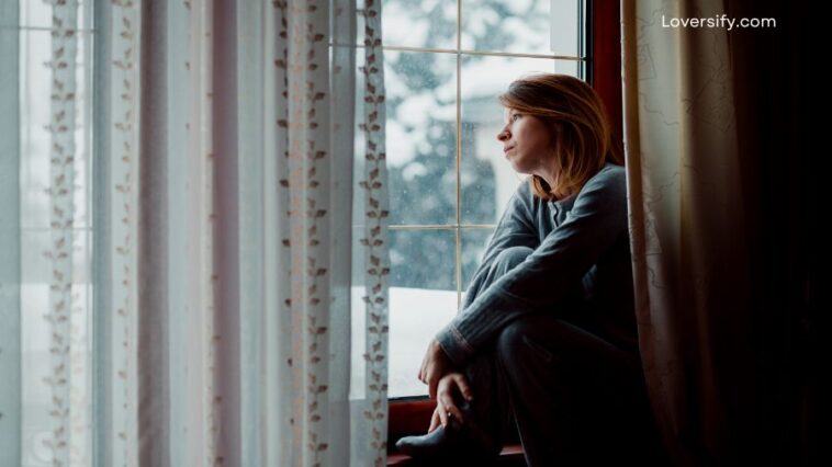 A woman in pajamas sits on a windowsill looking out a rain-streaked window.