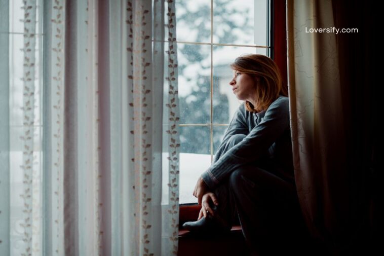 A woman in pajamas sits on a windowsill looking out a rain-streaked window.