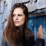A young woman with long brown hair leans against a weathered blue wooden door and wall, her hand resting on the door.