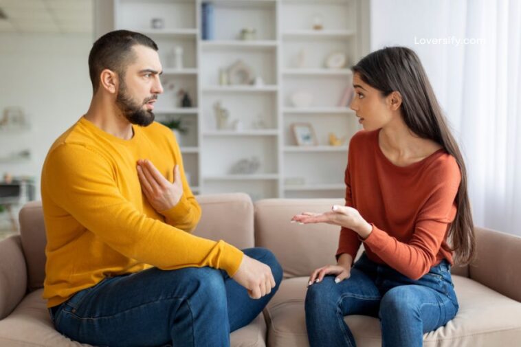 A man in a yellow sweater with his hand on his chest and a woman in an orange shirt with her hand outstretched are sitting on a couch and talking.