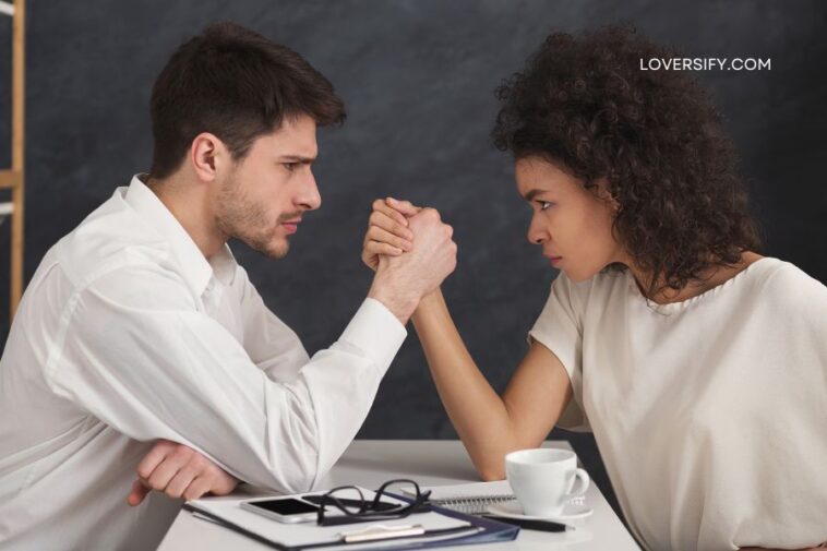Man and woman arm wrestling intently across a white table, with a coffee cup and notebook visible.