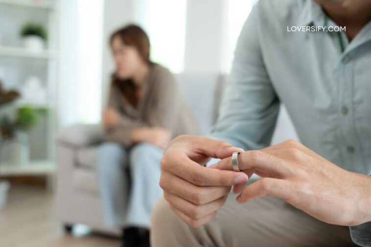 A man's hands remove a ring from his finger while a woman sits in the background, looking away.