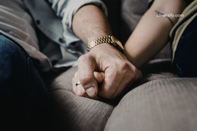 Two people holding hands, one wearing a gold watch and ring, sitting on a textured couch.