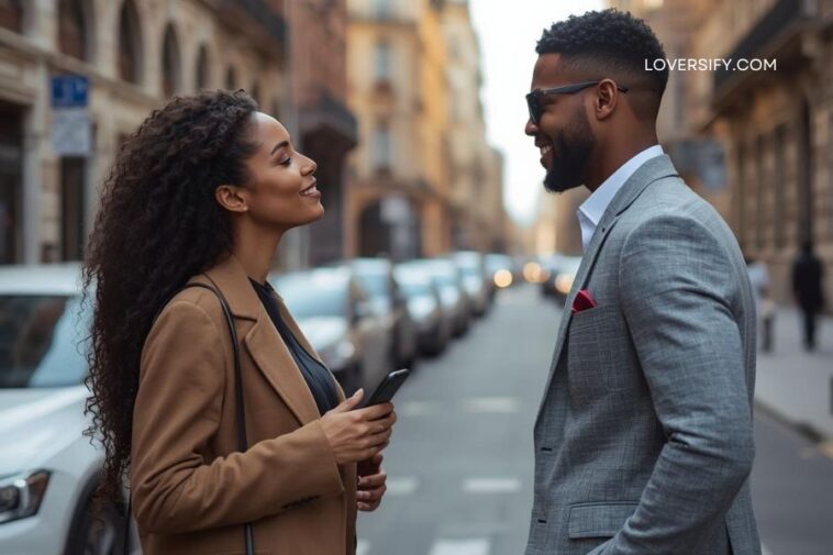 A man and a woman in stylish attire stand on a city street, facing each other and smiling. The woman holds a smartphone, and the man has a red pocket square. Cars are parked in the background along the street.