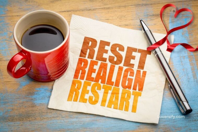 A red mug of coffee sits on a wooden surface next to a white napkin with the words "RESET REALIGN RESTART" printed in colorful letters. A silver pen rests on the napkin, and a red ribbon is shaped into a heart behind it.