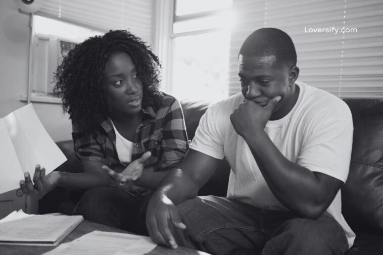 A Black woman gestures with papers while talking to a Black man who rests his chin on his hand, both seated on a couch in a sunlit room with papers on a coffee table.