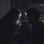 A woman and man in silhouette, facing each other and talking, are illuminated from the front in a dim room.