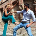 A blonde woman in a teal jumpsuit lunges forward with an outstretched arm and open mouth as a man in a blue patterned shirt and jeans playfully blocks her.