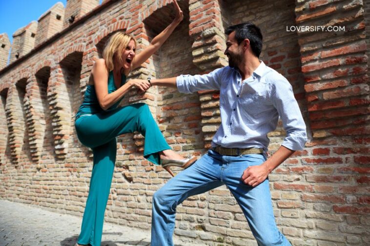 A blonde woman in a teal jumpsuit lunges forward with an outstretched arm and open mouth as a man in a blue patterned shirt and jeans playfully blocks her.
