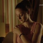 A woman in a pink top looks down with her hand on her shoulder in a dimly lit room with striped wallpaper.
