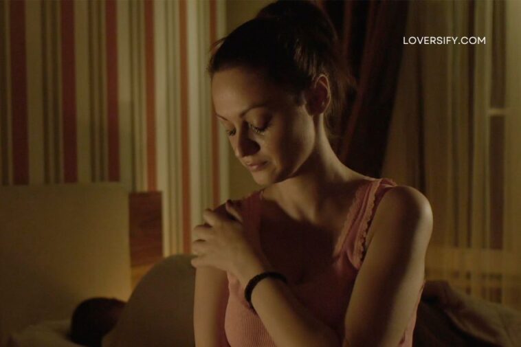 A woman in a pink top looks down with her hand on her shoulder in a dimly lit room with striped wallpaper.