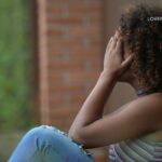 A woman with curly hair sits with her back against a wooden beam, her hands covering her face.