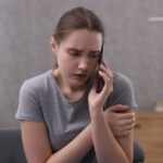 A young woman with a concerned expression speaks on a dark flip phone, holding it to her ear with her right hand and hugging her left arm with her right hand.