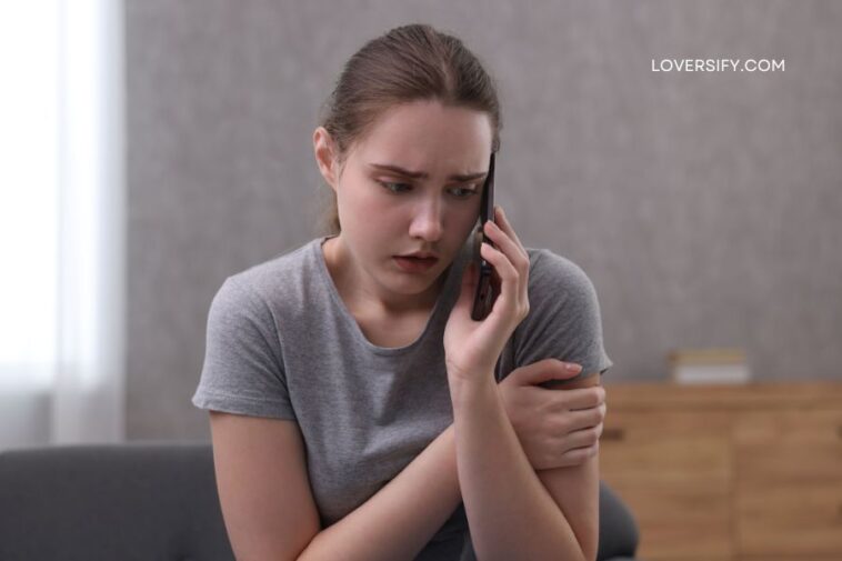 A young woman with a concerned expression speaks on a dark flip phone, holding it to her ear with her right hand and hugging her left arm with her right hand.