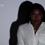 A young Black woman with dark hair and dark lipstick, wearing a white button-down shirt, looks directly at the camera. A large shadow of her head and shoulders is cast on the white wall behind her. The website "LOVERSIFY.COM" is visible in the upper left corner.