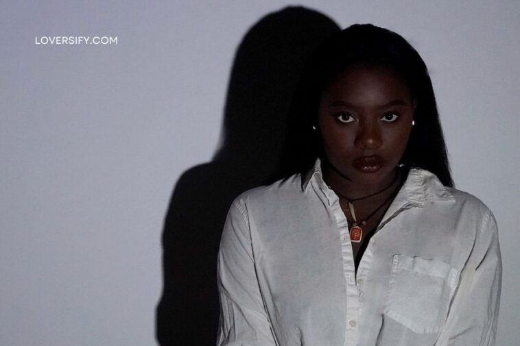 A young Black woman with dark hair and dark lipstick, wearing a white button-down shirt, looks directly at the camera. A large shadow of her head and shoulders is cast on the white wall behind her. The website "LOVERSIFY.COM" is visible in the upper left corner.