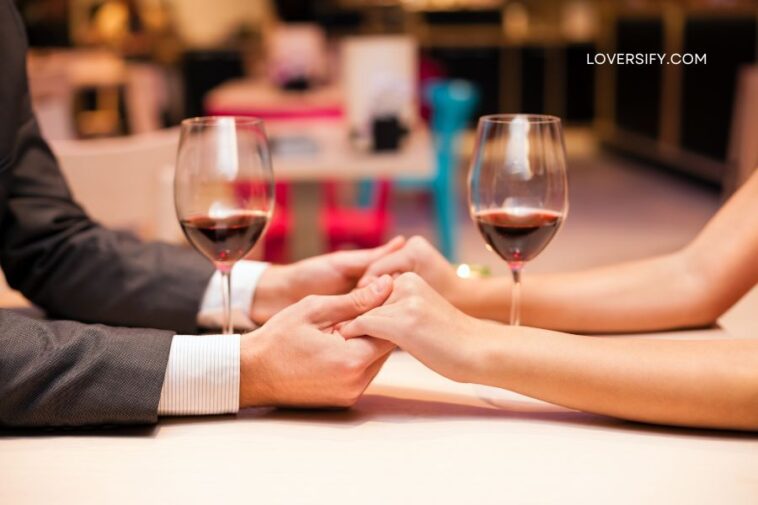 A couple's hands are clasped across a table with two glasses of red wine.