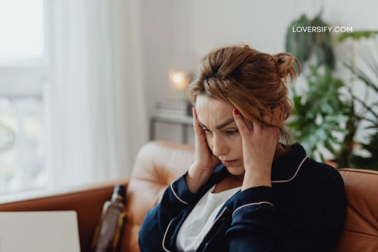 A woman with her hands on her temples sits on a brown leather couch, looking down with a distressed expression.