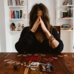 A woman with her hands clasped over her face sits at a wooden table, with a photo and jewelry spread before her.