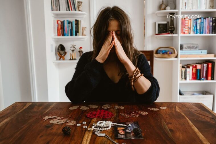 A woman with her hands clasped over her face sits at a wooden table, with a photo and jewelry spread before her.