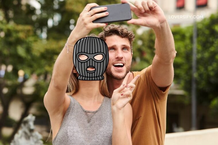 A man and woman take a selfie outdoors, the woman's face is covered by a black balaclava graphic, and the man holds up two fingers in a peace sign.