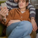 A young couple embraces on the floor with a green sofa behind them; the woman holds a smartphone.