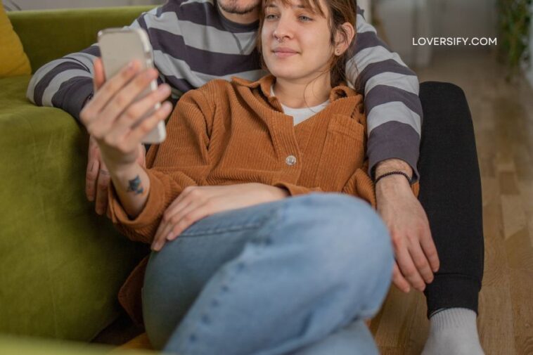 A young couple embraces on the floor with a green sofa behind them; the woman holds a smartphone.