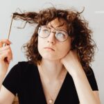 Young woman with curly brown hair and glasses, wearing a black shirt, holds a golden pen to her hair and rests her chin on her hand while looking away.