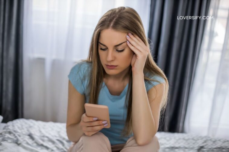 A young woman with light brown hair sits on a bed, holding a light pink smartphone and touching her forehead with her other hand.
