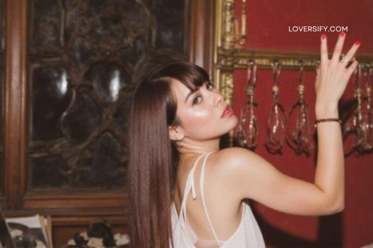 A woman with long brown hair and bangs, wearing a white camisole, looks over her shoulder towards the camera, her right arm raised. She is in a dimly lit room with ornate dark wood furniture on the left and a red wall with golden trim and hanging glass objects on the right.