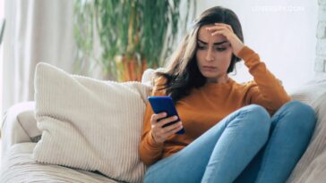 A young woman with dark hair sits on a couch, holding a blue smartphone and touching her forehead with her hand.