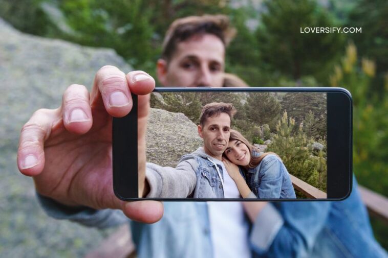 Man holding a smartphone and taking a selfie of himself and a woman, with a rocky, wooded background.