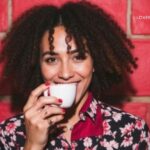 A smiling woman with curly hair drinks from a white teacup against a red brick wall.