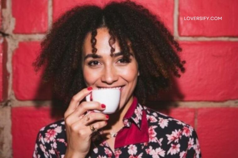 A smiling woman with curly hair drinks from a white teacup against a red brick wall.