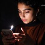 A young person with dark curly hair holds a smartphone in a dark room, illuminated by the phone's bright screen and the flash of a camera or light source.
