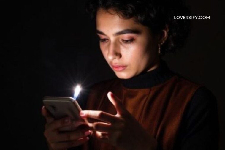 A young person with dark curly hair holds a smartphone in a dark room, illuminated by the phone's bright screen and the flash of a camera or light source.