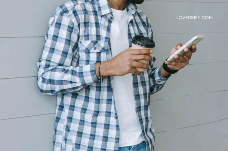 A person in a blue plaid shirt and white t-shirt holds a coffee cup and a smartphone while leaning against a gray wall.