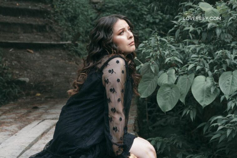 A woman with long, dark, wavy hair wearing a black lace dress sits on a wooden structure next to lush green foliage.