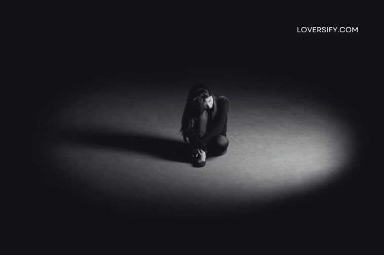 A solitary woman with long, dark hair crouches on a dark, textured floor, illuminated by a single spotlight. She is curled inward, with her knees pulled to her chest and her head bowed. The background is a deep, dark void, with only a faint glow from the spotlight to define the space.