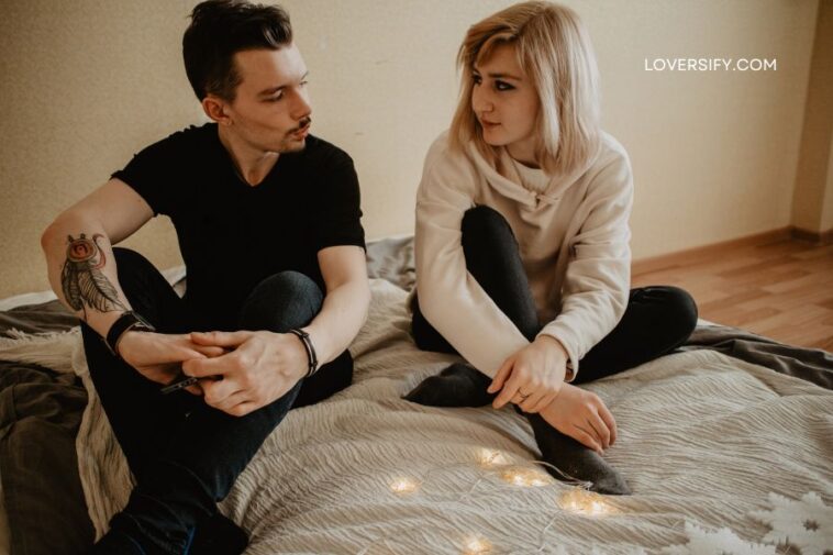 A man and a woman sit on a bed, looking at each other, with a string of fairy lights between them.