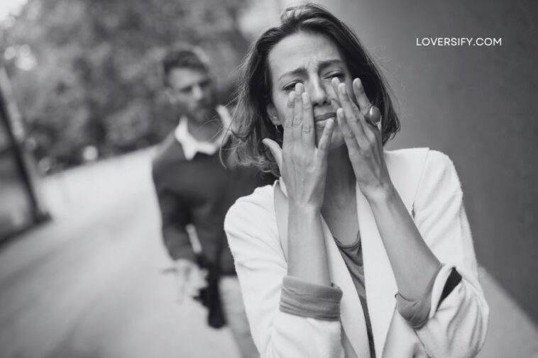 A woman cries, covering her face with her hands, while a man walks away in the background.