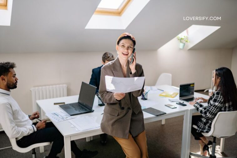 A woman in a brown blazer talks on the phone while holding documents in an office setting with three other people visible.