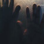 Silhouetted hands press against a frosted glass surface, illuminated by warm light from behind.