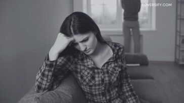 A woman in a plaid shirt rests her forehead on her hand, looking down with a sad expression, while a man stands with his back to the camera in front of a window in the background.