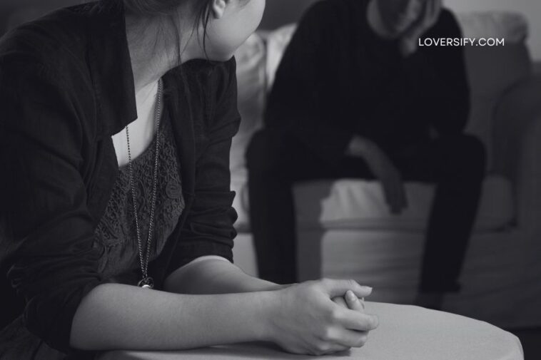 A woman in the foreground with her hands clasped sits facing away from the camera, while a man in the background, blurred and slumped on a sofa, holds his head in his hand.