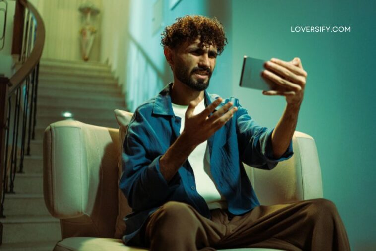 Man with curly hair sitting on a sofa, holding a tablet and gesturing with his hands. A staircase is visible in the background.
