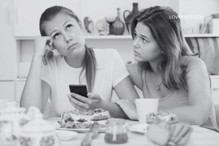 Two young women sit at a table with food, one holding a phone with a bored expression, the other looking at her with concern and placing a hand on her arm.