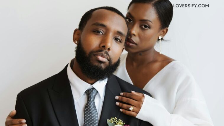 A Black man in a suit and tie, with a boutonnière, is posed with a Black woman in a white dress, her arm around his shoulder, against a plain background.